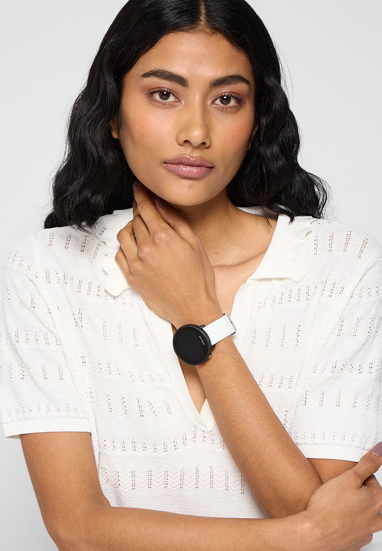 Reloj inteligente negro redondo con una correa de silicona blanca, exhibido en una muñeca. Suéter blanco de punto texturizado de fondo.