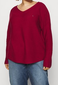 Pull en V bordeaux à manches longues et à la coupe décontractée. Fabriqué en tissu tricoté texturé, avec un petit logo près de la poitrine.