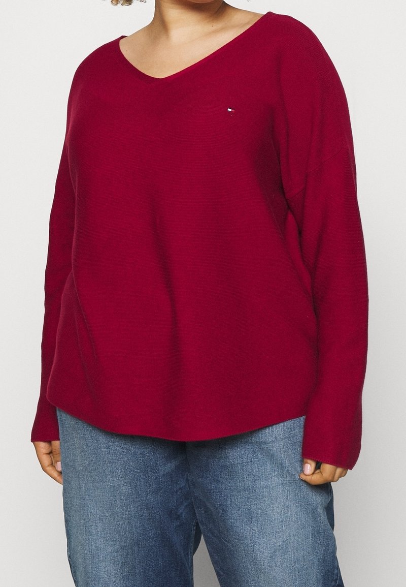 Pull en V bordeaux à manches longues et à la coupe décontractée. Fabriqué en tissu tricoté texturé, avec un petit logo près de la poitrine.