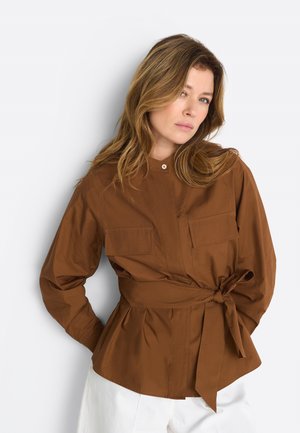 Heledate juustega naine, kellel on pruun vööga jakk taskutega ja valged püksid, seisab ühevärvilise heleda tausta ees.