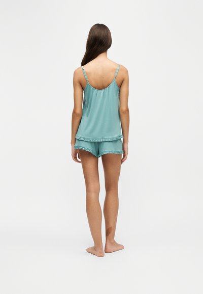 Femme aux longs cheveux bruns, debout pieds nus, de dos, portant un haut bleu clair sans manches et un short assorti avec un bord volanté.