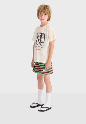 Dreng står iført en cremefarvet T-shirt med sort-hvid hundetryk, grønne og vinrøde stribede shorts, hvide sokker og sorte sandaler.