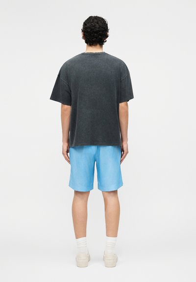 Homme aux cheveux bouclés vu de dos, portant un t-shirt gris foncé ample, un short bleu clair, des chaussettes blanches et des baskets beiges.