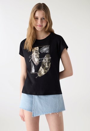 Giovane donna che indossa una maglietta nera con motivo di piuma dorata e una gonna di jeans asimmetrica azzurro chiaro su sfondo neutro.