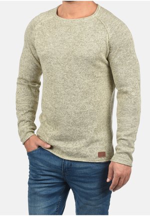 Mann trägt beige langärmeliges, gestricktes Sweatshirt und blaue Jeans, eine Hand in der Tasche, neutraler Hintergrund.