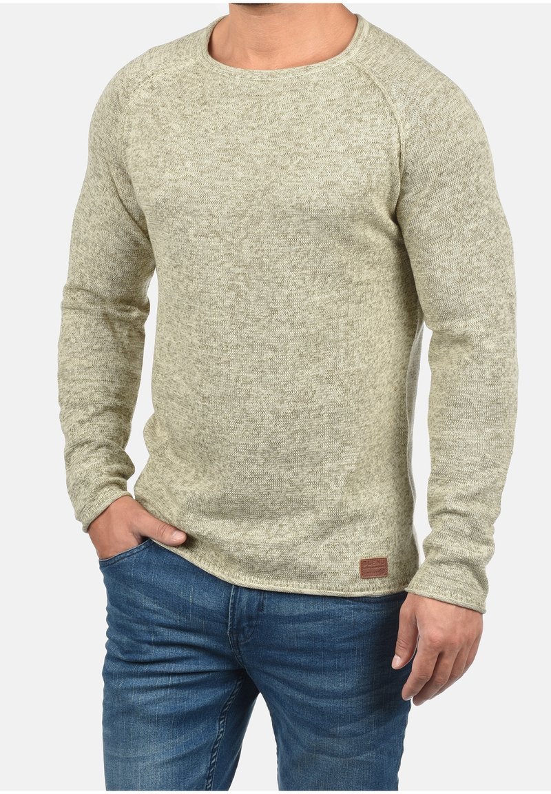 Homme portant un pull en laine beige à manches longues et un jean bleu, une main dans la poche, sur un fond neutre.