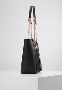 Musta teksturoidusta nahasta valmistettu käsilaukku, jossa on kultaketjuolkkaat ja ruusukultaiset detaljit, seisoo pystyasennossa valkoisella jalustalla tasaisen taustan edessä.