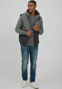 Grauer Kapuzenwesten über einem grauen Langarmshirt, blaue Jeans und weiße Sneaker. Die Weste hat eine strukturierte Oberseite und Reißverschlusstaschen.