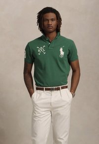 Ung mann med dreads iført en grønn polo med hvite logoer og beige bukser, stående med hendene i lommene foran en nøytral bakgrunn.