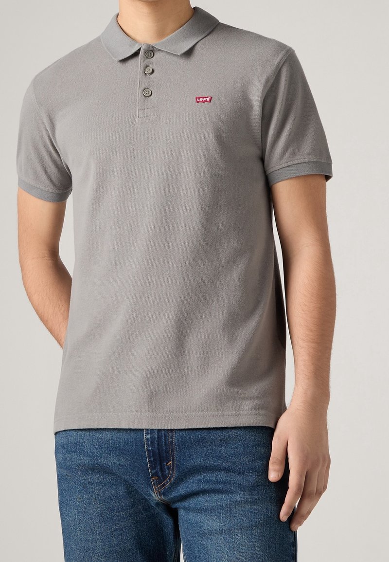 Homme portant un polo gris à manches courtes avec trois boutons et un petit logo rouge Levi's, associé à un jean bleu.