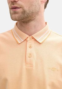 Polo shirt orange clair avec un tissu texturé, deux boutons et un col arborant des rayures blanches et orange. Logo notable sur la poitrine.