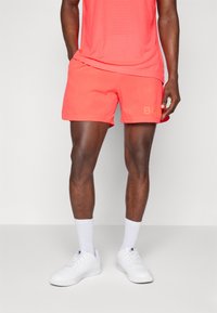 Coral athletic shorts with a soft texture and elastic waistband, paired with white sneakers and crew-length socks. Logo detail on shorts.