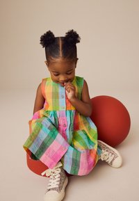 Kolorowa, w paski, bezrękawnikowa sukienka z guzikami i marszczonym dołem. Dziecko w błyszczących trampkach, siedzące obok czerwonej piłki.