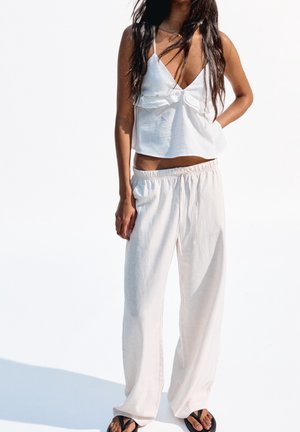 Weißes, kurzes Tanktop mit Rüschensaum, kombiniert mit lockeren, hellbeigen Hosen mit elastischem Bund und Kordelzug. Schwarze Flip-Flops.