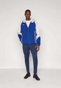 Veste coupe-vent bleue et blanche avec capuche, dotée d'une fermeture éclair et de poches latérales, associée à un jogging bleu marine et des chaussures de sport noires.
