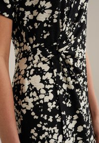 Černé šaty s bílým květinovým vzorem. Mají texturovanou látku a uzlový detail v pase. Detailní pohled z boku.