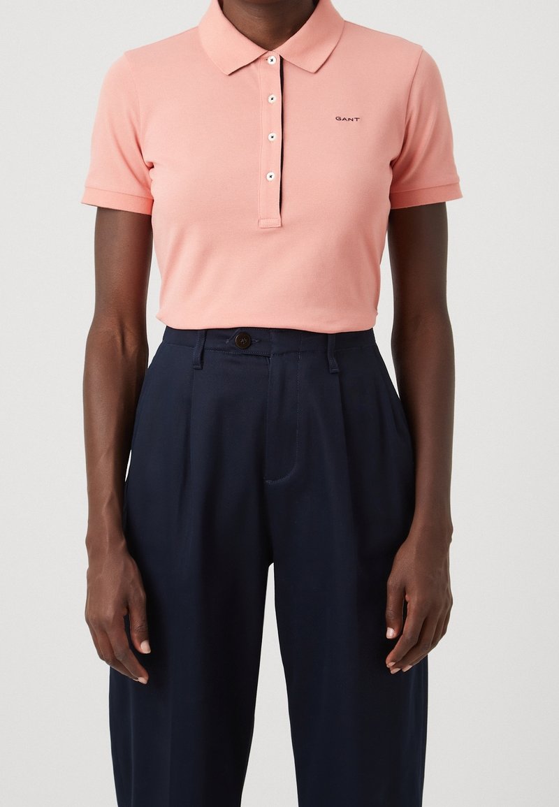 Osoba ubrana w łososiowy różowy krótki rękaw polo, włożony w granatowe spodnie z wysokim stanem i plisami.