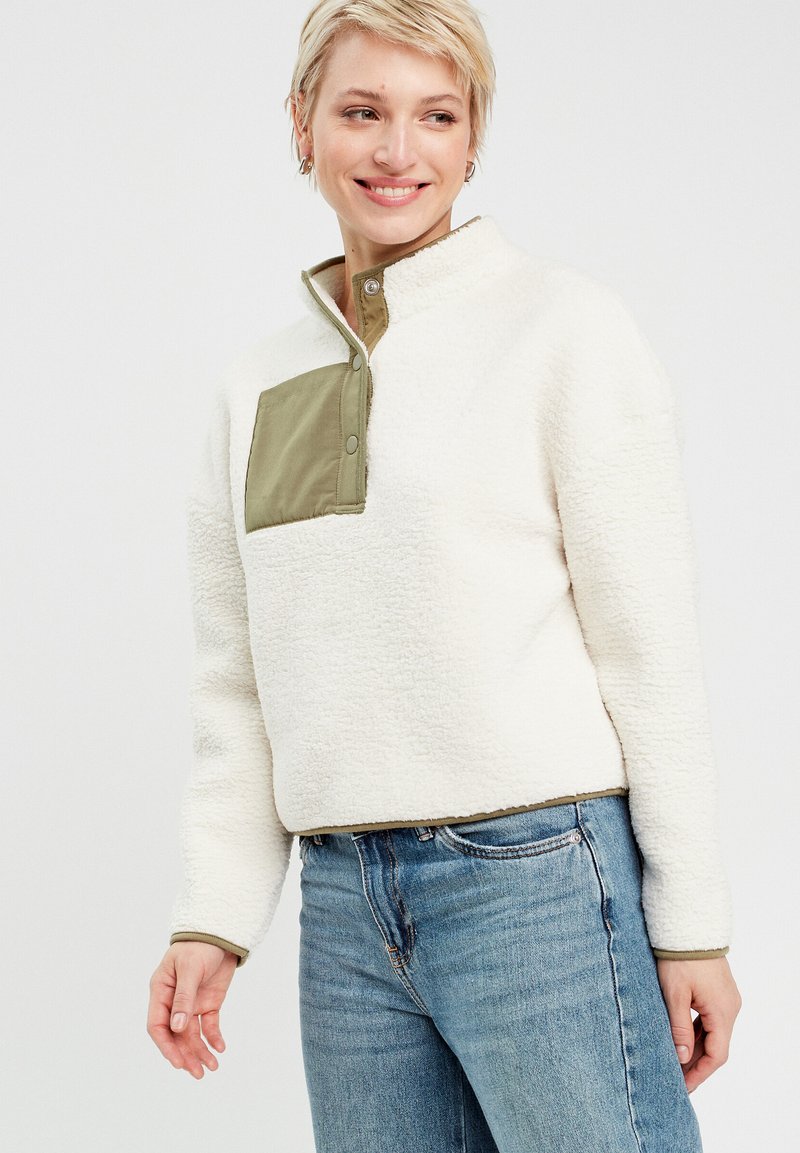 Weißer Fleece-Pullover mit einer grün geknöpften Brusttasche und Kragen. Verfügt über eine weiche, strukturierte Oberfläche und ein cropped Design. Getragen mit blauen Jeans.