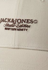 Beige tygkeps med brunt broderat text som står "JACK & JONES Studio Edition NINETEEN NINETY" på frampanelen.