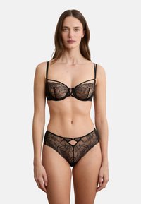 Frau trägt schwarzen Spitzen-BH und passende Spitzen-Höschen mit hohem Bund, steht frontal vor weißem Hintergrund.