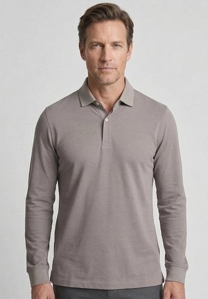 Mann mit kurzen braunen Haaren, der ein graues Langarm-Poloshirt trägt, steht vor einem schlichten hellgrauen Hintergrund.