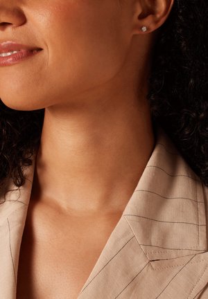 Zilveren studoorbel op een model met krullend haar, die een lichtbeige, gestreepte blazer draagt. De huidtoon lijkt warm en heeft een gladde textuur.