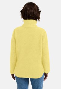 Pullover in pile giallo con colletto alto, esterno testurizzato e spalle scese, abbinato a jeans in denim blu. Vista posteriore mostrata.