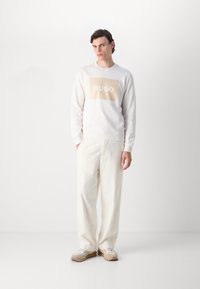 Weißer Sweatshirt mit einem beigen rechteckigen Logo, das "HUGO" in Weiß zeigt. Kombiniert mit lockeren cremefarbenen Hosen und hellgrauen Sneakers.