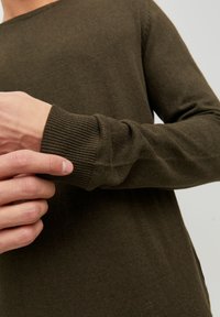 Sötét olívazöld kötött, hosszú ujjú pulóver ribbelt mandzsettákkal. Közeli kép egy kézről, amely a hajat állítja be, megmutatva a textúrát és az anyag részleteit.