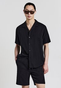 Maglietta nera a maniche corte con un motivo a rombi, con chiusura a bottoni, abbinata a pantaloni corti dello stesso tessuto testurizzato. Occhiali da sole indossati.