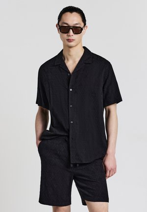 Schwarzes kurzärmeliges Hemd mit einem Rautenmuster, Knopfschluss, kombiniert mit passenden Shorts aus demselben strukturierten Stoff. Sonnenbrille getragen.