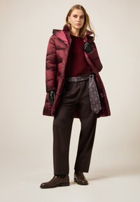 Bordowy puchowy płaszcz z wysokim kołnierzem, zestawiony z bordowym swetrem, brązowymi spodniami i brązowymi skórzanymi botkami za kostkę. Pasek z wzorem.