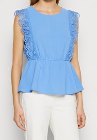 Haut péplum bleu clair avec finition en dentelle sur les épaules. Confectionné dans un tissu doux, présentant un col rond et une taille élastiquée pour une coupe décontractée.