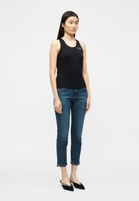 Γυναίκα που στέκεται, φοράει μαύρο αμάνικο τοπ με λογότυπο, τζιν παντελόνι καμπάνα μέχρι τον αστράγαλο και μαύρες μυτερές μπαλαρίν ες, μπροστά σε απλό λευκό φόντο.