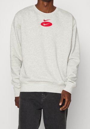 Man wearing light gray sweatshirt with red double Nike swoosh logo on chest and dark gray jeans, standing against plain background.