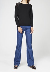 Musta pitkähihainen neulepaita, jossa pitsikoristelu olkapäillä, yhdistetty liehuhousuihin ja mustiin loafer-kenkiin. Yksinkertainen ja istuva malli.