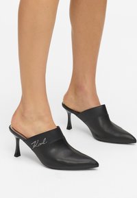 Czarne skórzane klapki z wąskimi noskami i niskim obcasem w kształcie szpilki. Charakteryzują się eleganckim designem oraz tłoczonym logo na górnym pasku.