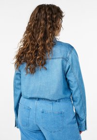 Osoba s kučeravými vlasmi, ktorá má na sebe svetlomodrú rifľovú bundu a džínsy v rovnakom odtieni, zobrazená zozadu na bielom pozadí.