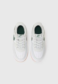 Hvite og grå Nike-sneakers med grønne detaljer, glatt læroverdel, perforert tåboks og gummisåle. Har snørebånd og logo.