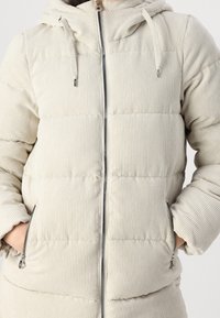 Beige puffer-jakke med vertikal ribbet struktur, hætte, frontlynlås og sidelommer. De fyldte sektioner er syet i vandrette linjer.