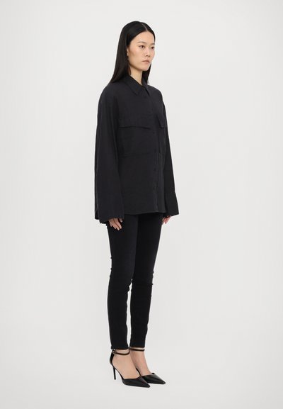 Black long-sleeve shirt with two chest pockets, paired with black skinny jeans and pointed-toe heels, creating a streamlined silhouette.