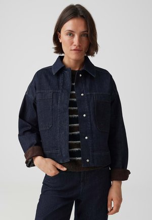 Frau mit kurzen braunen Haaren trägt eine dunkle Jeansjacke über einem gestreiften Pullover und dunklen Jeans, posiert mit einer Hand in der Tasche.