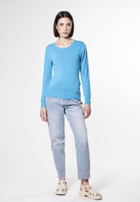 Maglione blu a maniche lunghe con scollatura rotonda, texture morbida, abbinato a jeans chiari leggermente strappati e sandali beige.