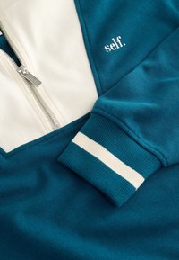 Sweat-shirt à fermeture éclair bleu et blanc avec "self." brodé sur la poitrine et une bande blanche sur le bord de la manche.