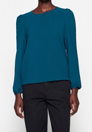 Person wearing a long-sleeve teal blouse with puffed shoulders and elastic cuffs paired with black pants.