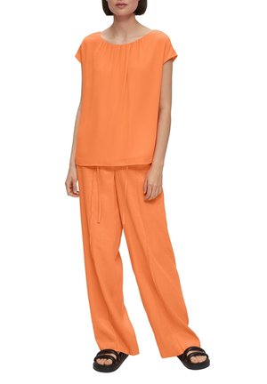 Orange, kurzärmliges Oberteil mit gerafftem Ausschnitt, kombiniert mit lockeren, hochgeschnittenen Hosen. Strukturierter Stoff; flache, schwarze Sandalen.
