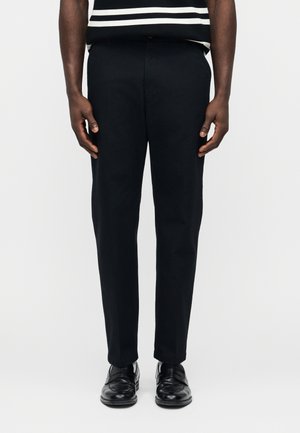 Άνδρας που φοράει μαύρο παντελόνι ραμμένο κατά παραγγελία, μαύρα δερμάτινα παπούτσια και πουκάμισο με μαύρες και άσπρες ρίγες, στέκεται μπροστά σε απλό φόντο.