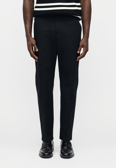 Homme portant un pantalon noir sur mesure, des chaussures en cuir noires et une chemise à rayures noires et blanches, debout devant un fond uni.