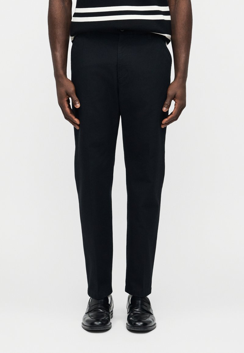 Άνδρας που φοράει μαύρο παντελόνι ραμμένο κατά παραγγελία, μαύρα δερμάτινα παπούτσια και πουκάμισο με μαύρες και άσπρες ρίγες, στέκεται μπροστά σε απλό φόντο.