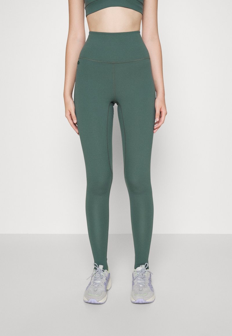 Leggings taille haute de couleur vert foncé, fabriqués en tissu extensible, avec des coutures plates. Associés à des baskets gris clair avec des accents violets.